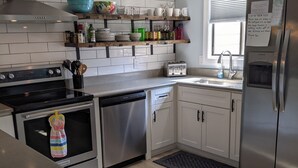Een moderne keuken met witte kasten, een roestvrijstalen koelkast, een vaatwasser en een fornuis. Boven de aanrecht staan planken met diverse keukenbenodigdheden.