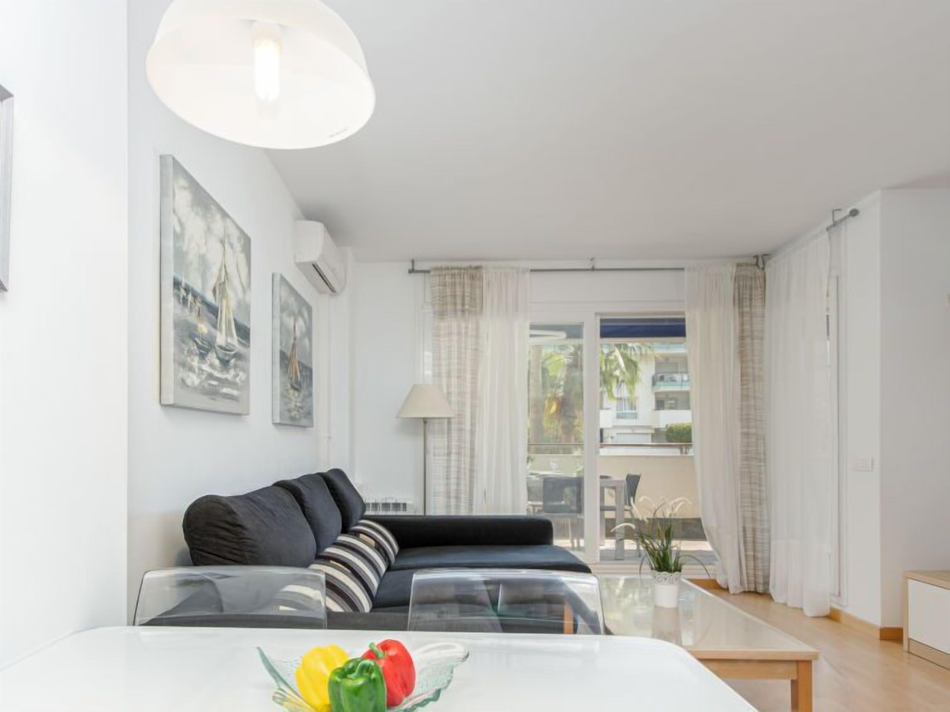 Una sala de estar moderna con un sofá, una mesa de centro de vidrio con fruta, y vistas a través de ventanas amplias.