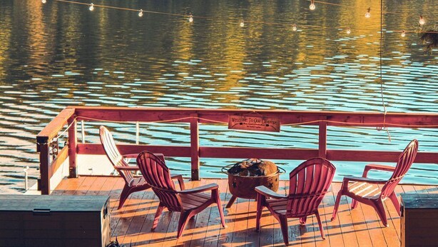 Una terraza de madera con cuatro sillas Adirondack, un brasero de metal y luces colgantes sobre el agua.