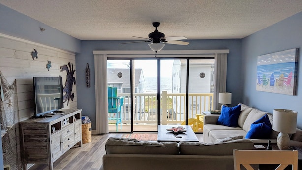Ein Wohnzimmer mit einer Couch, einem Couchtisch, einem Fernseher und einem Balkon mit Meerblick.
