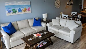 Ein Wohnzimmer mit zwei weißen Sofas, einem Holztisch, einem Deckenventilator, einem Bild von Strandstühlen und einem Essbereich mit weißen Stühlen und einem Tisch.