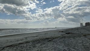 Ein Strand mit einem Steg, der sich ins Meer erstreckt, feiner Sand und Gebäude im Hintergrund.