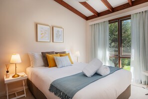 Quarto com uma cama grande, duas mesas de cabeceira, janela com cortinas e duas gravuras de plantas emolduradas na parede.