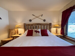 Una cama con cabecera de madera, dos mesitas de noche con lámparas, y una ventana con cortinas rojas.