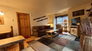 A cozy living room with a wooden door, a sofa, a coffee table, and a television. - Moose Ski Condo in Canyons Village of Park City 3-minutes from Slopes & Trails! (Park City)