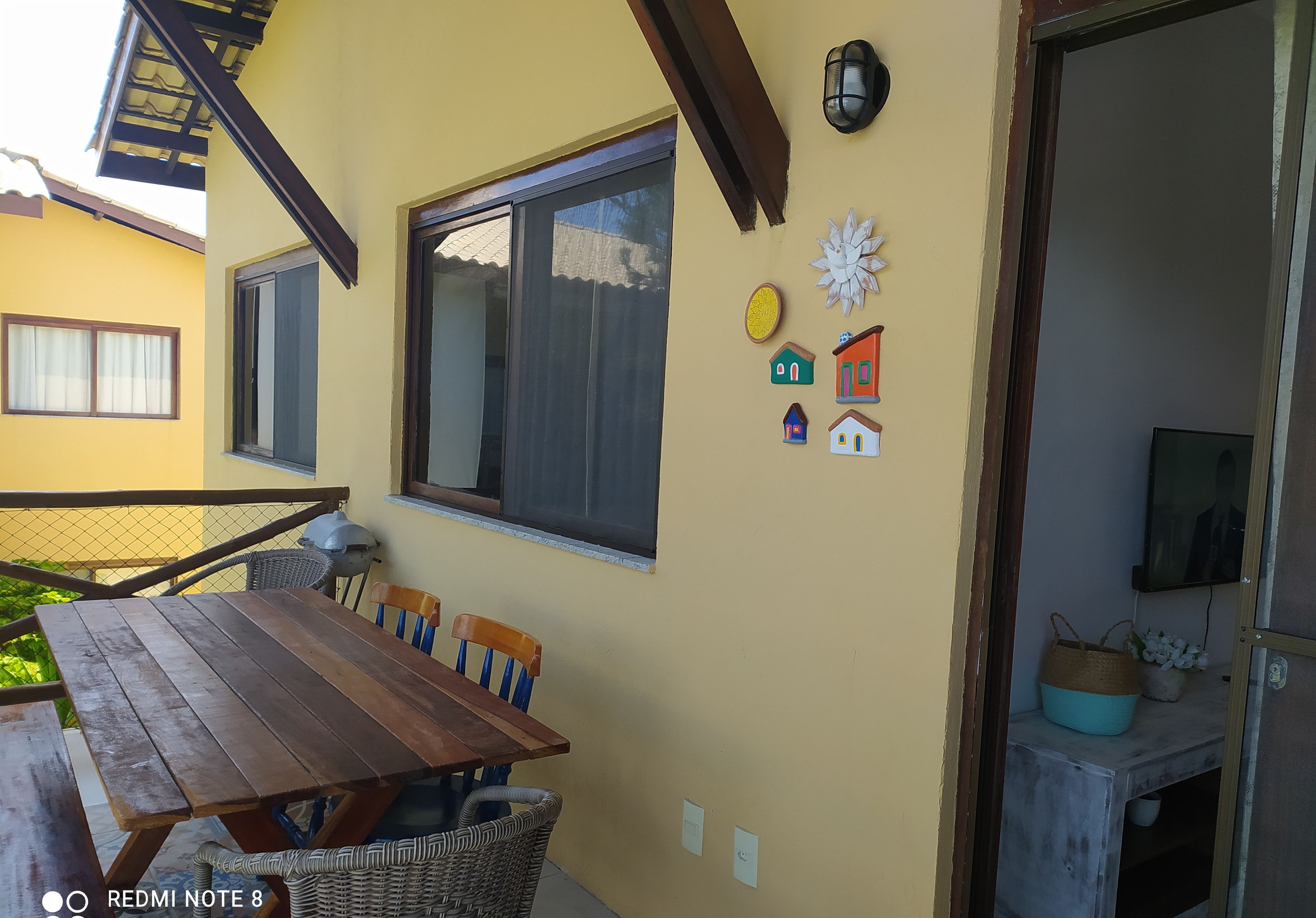 Un comedor con mesa de madera y sillas de mimbre, ventana con cortina y decoración en la pared, incluyendo un sol y casas.