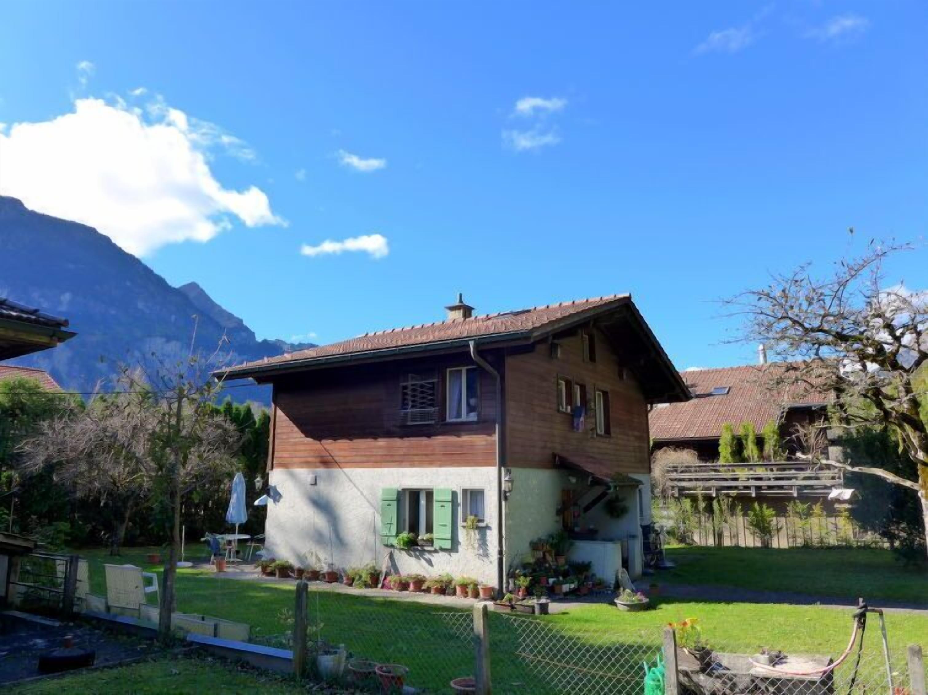 Une maison en bois à deux étages, dotée d’un toit en pente, entourée d’une cour clôturée où se trouvent des plantes en pot, avec une montagne en arrière-plan.