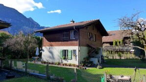 A two-story wooden house with a sloped roof, surrounded by a fenced yard with potted plants and a mountain in the background.