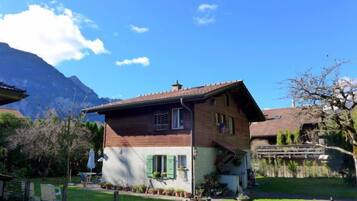 Une maison en bois à deux étages, dotée d’un toit en pente, entourée d’une cour clôturée où se trouvent des plantes en pot, avec une montagne en arrière-plan.