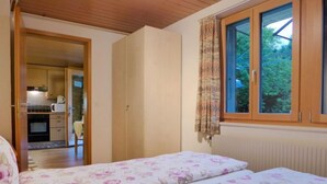 A bedroom with two beds, a window with curtains, a view of the outdoors, and a kitchen in the adjacent room.