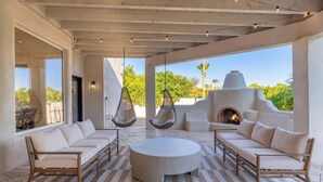 Un vaste espace de détente extérieur comprenant une table ronde, des canapés blancs, des fauteuils suspendus et une cheminée.
