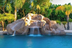 A pool with a rock waterfall feature surrounded by lush greenery.