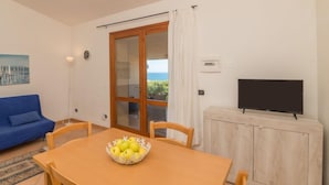 Ein Wohnzimmer mit einer Couch, einem Esstisch mit Obstschale, einem Fernseher und einer Glastür zum Balkon.