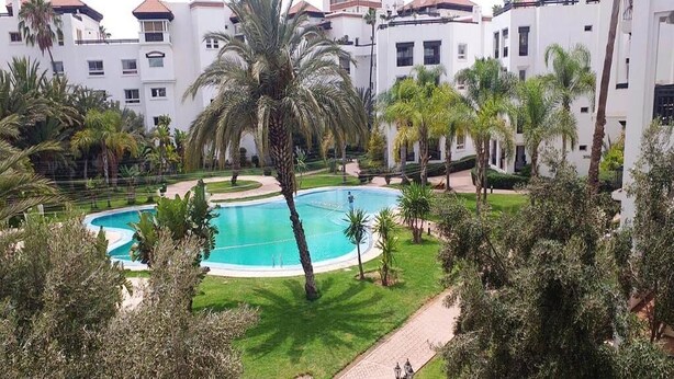 Ein Pool, umgeben von Palmen, und ein Weg, der zu einem mehrstöckigen Gebäude mit Balkonen führt.