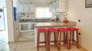 A kitchen with white cabinets, a granite countertop, and a window with blinds. There are three wooden stools with red cushions. - Summer Center Imi Ouaddar 5389 (Imi Ouaddar)