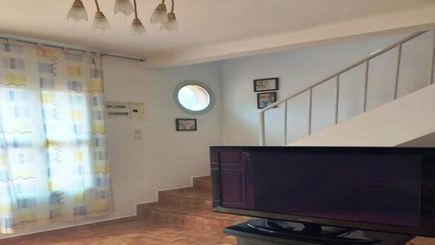 Una sala de estar con un televisor de pantalla plana sobre un mueble bajo, una escalera y un ventanal con cortinas.