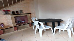 Un comedor con una mesa redonda y cuatro sillas blancas, una chimenea de piedra con adornos, y una escalera con barandilla blanca.