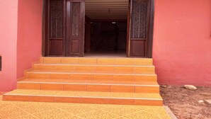 Un escalier en bois mène à une porte en bois sombre ornée de motifs complexes, le tout se détachant sur un mur rose.