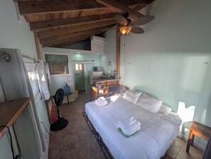 A bedroom with a bed, a ceiling fan, a small table, and a kitchenette in the background. - Beach Front! Cobia Beach Guest House, Pink and Teal together (Hopkins)