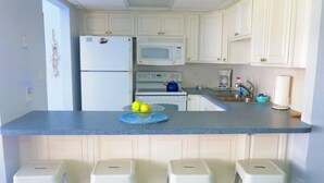 Una cocina con armarios blancos, encimera azul, refrigerador blanco, microondas, hervidor y tres taburetes blancos.