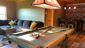 Una mesa de billar en una habitación con techo de madera, pared de ladrillo y un sofá con cojines.