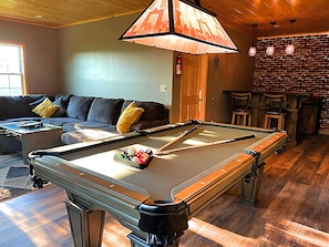 Una mesa de billar en una habitación con techo de madera, pared de ladrillo y un sofá con cojines.