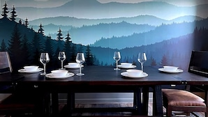 Una mesa de comedor dispuesta con copas de vino y platos, con una vista panorámica de montañas al fondo.
