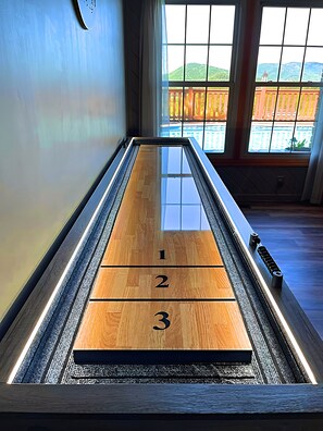 Uma mesa de jogo de shuffleboard com áreas de pontuação numeradas, com vista para uma piscina e montanhas através de uma janela.