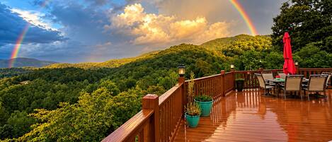 A wooden deck with outdoor furniture and potted plants, overlooking a forested landscape with a rainbow in the sky.