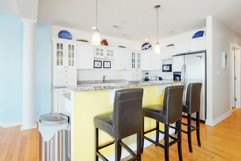 A modern kitchen with white cabinets, a granite countertop, and a stainless steel refrigerator. There are three bar stools and a trash can.