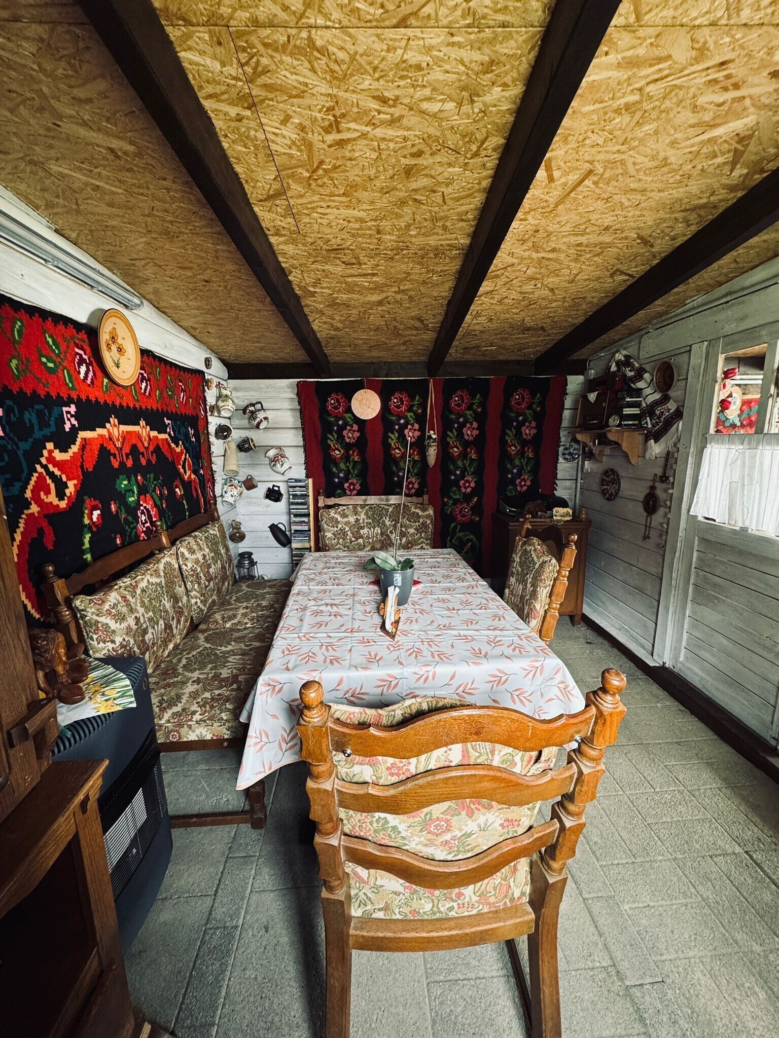 Een gezellige kamer met een houten plafond, een kamerkleed met dessin, een bank, een eettafel met stoelen en diverse decoratieve items aan de wanden.