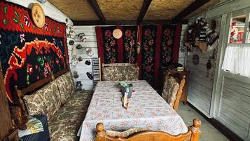 Een gezellige kamer met een houten plafond, een kamerkleed met dessin, een bank, een eettafel met stoelen en diverse decoratieve items aan de wanden.