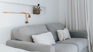 A grey sofa with two white cushions, a woven plant hanger, and a white wall. - Studio On Lewis Luxury Tiny House 4 mins to town (Mudgee)