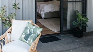 A wicker chair with a cushion, a hanging plant basket, and a potted plant on a balcony with a sliding glass door leading to a bedroom. - Studio On Lewis Luxury Tiny House 4 mins to town (Mudgee)