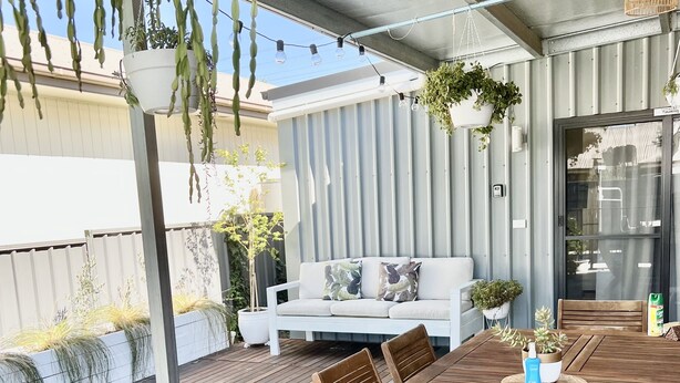 A wooden deck with a dining table and chairs, a white sofa with cushions, hanging plants, and a wicker light fixture. - Studio On Lewis Luxury Tiny House 4 mins to town (Mudgee)