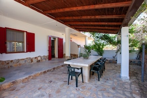 Une terrasse extérieure couverte, dotée d’un sol en pierre, d’une longue table avec des chaises et de volets rouges sur les murs.