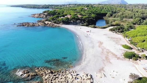 Une plage côtière aux eaux turquoise et limpides, parsemée de rochers et bordée d’une combinaison de zones naturelles et résidentielles.