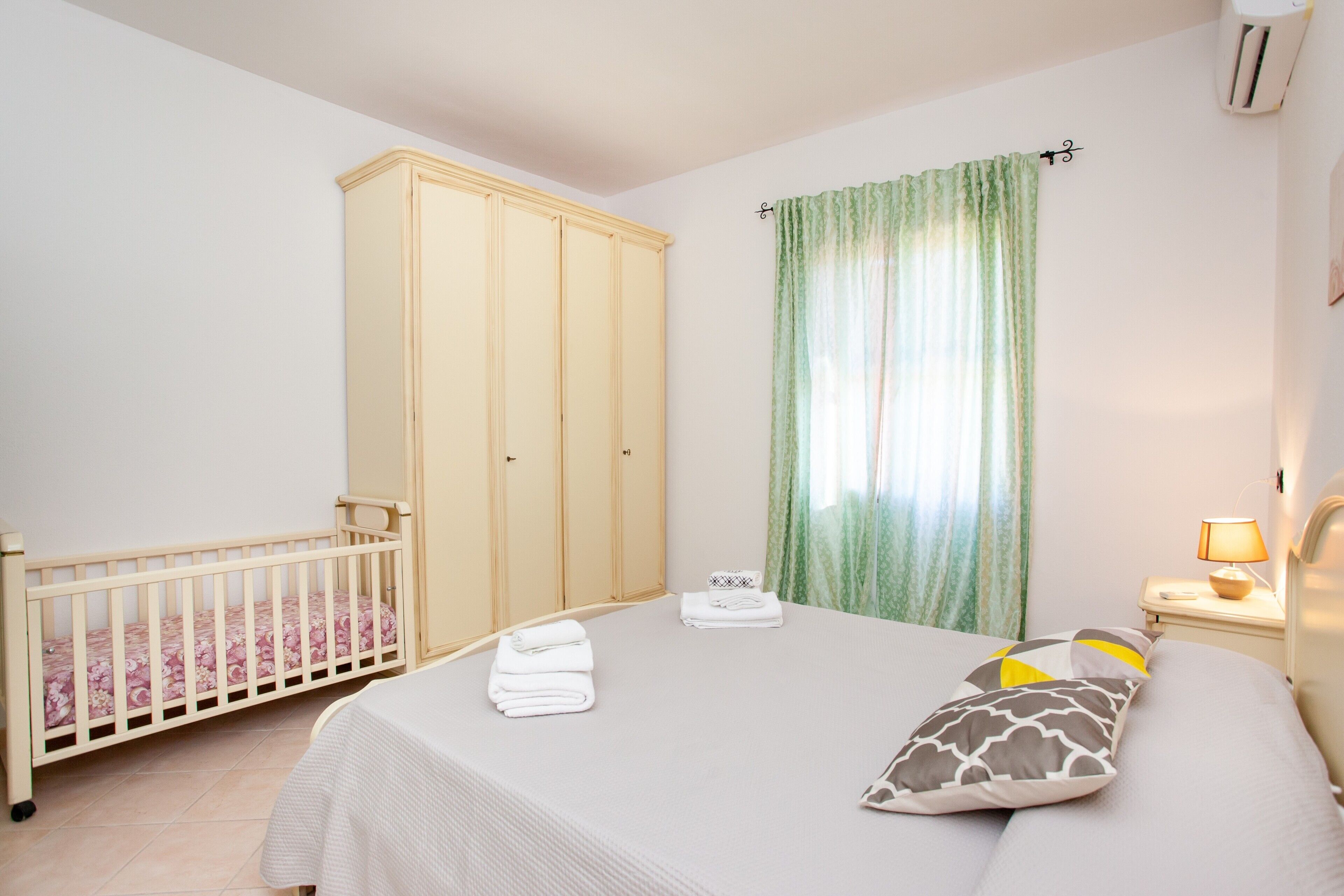 A bedroom with a crib, a bed with pillows, a wardrobe, and a nightstand with a lamp.