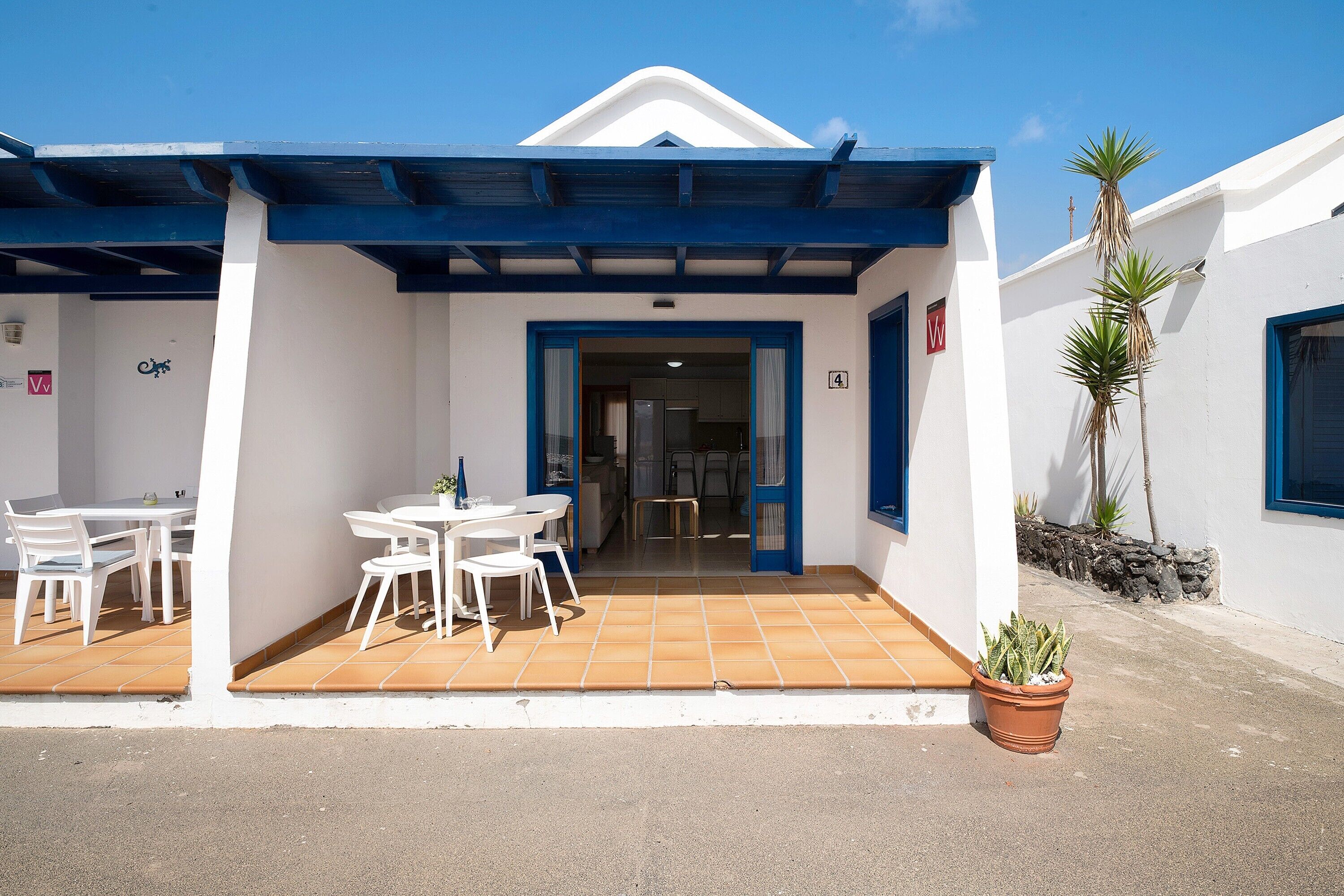 Un espace extérieur abrité, aménagé avec des tables et des chaises, un pot de fleurs et l’entrée d’un bâtiment.