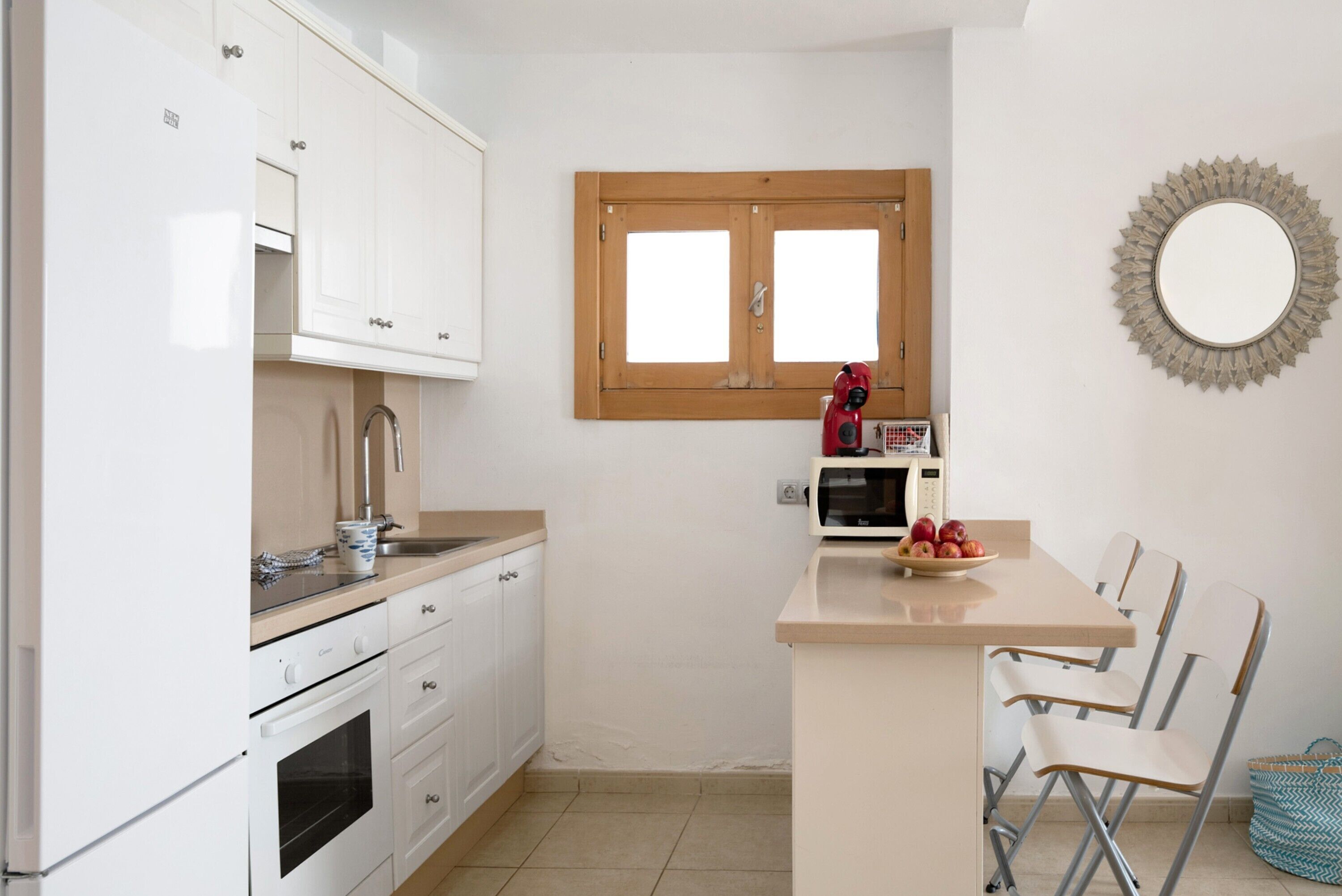 Cocina compacta con armarios blancos, microondas, pileta y un pequeño comedor con sillas.