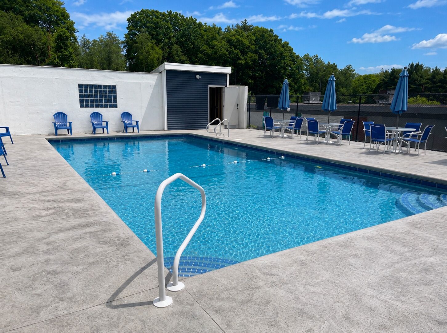 Ein rechteckiger Swimmingpool mit Metallleiter, umgeben von blauen Stühlen und Sonnenschirmen, und ein überdachter Bereich mit einer Tür.