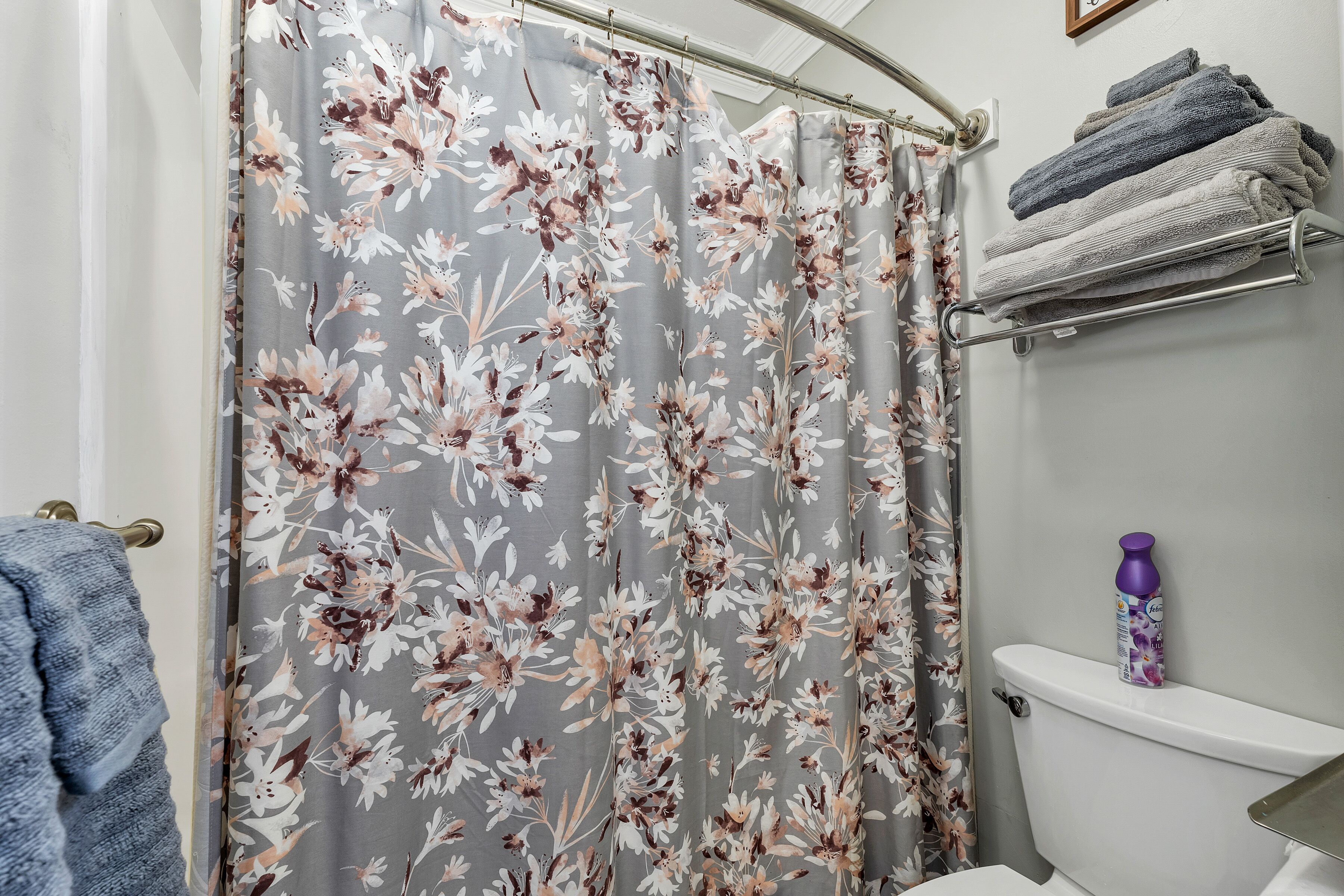 Un baño con cortina de ducha estampada con motivos florales, un perchero con toallas y un inodoro con una botella encima.