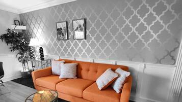 Un salon avec un papier peint à motifs, un canapé agrémenté de coussins à motifs, une table basse en verre et une table d’appoint avec une lampe.