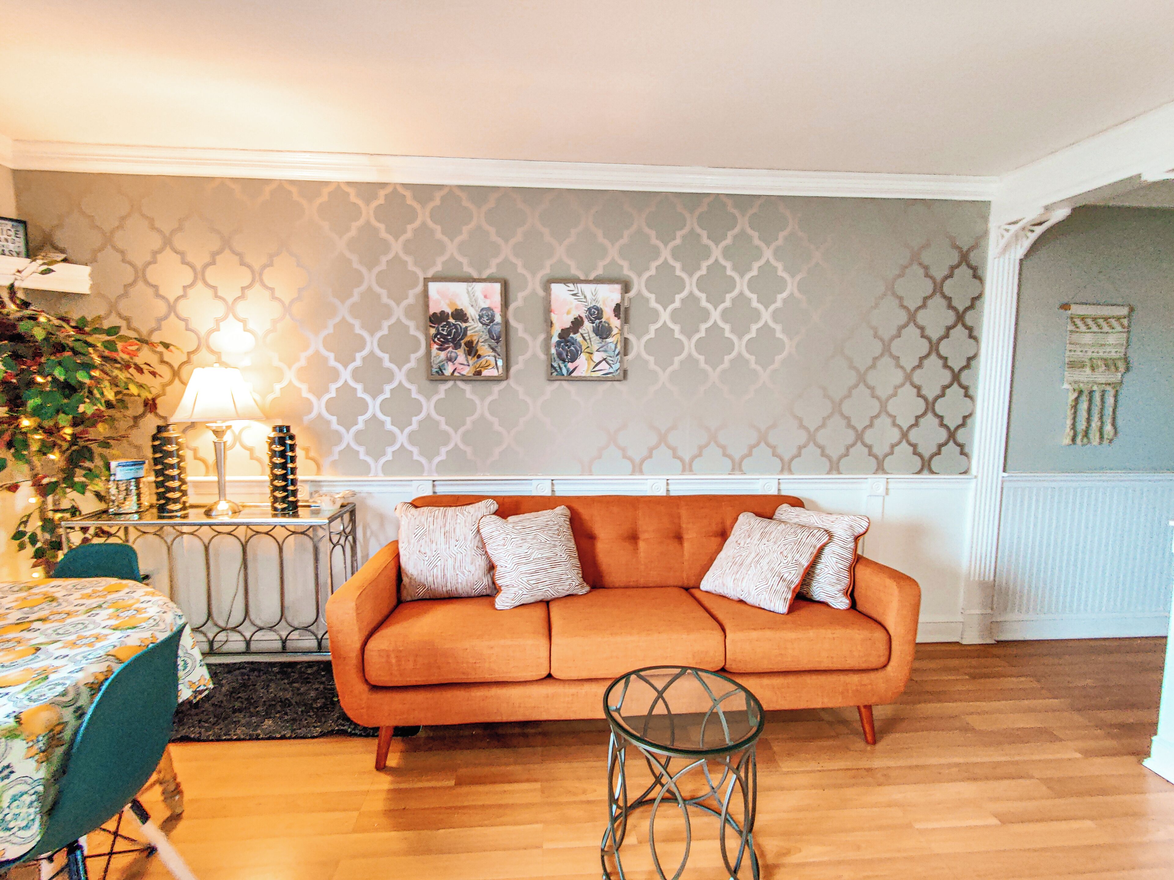A living room with a sofa, a glass coffee table, a patterned wall, and a small table with a lamp.