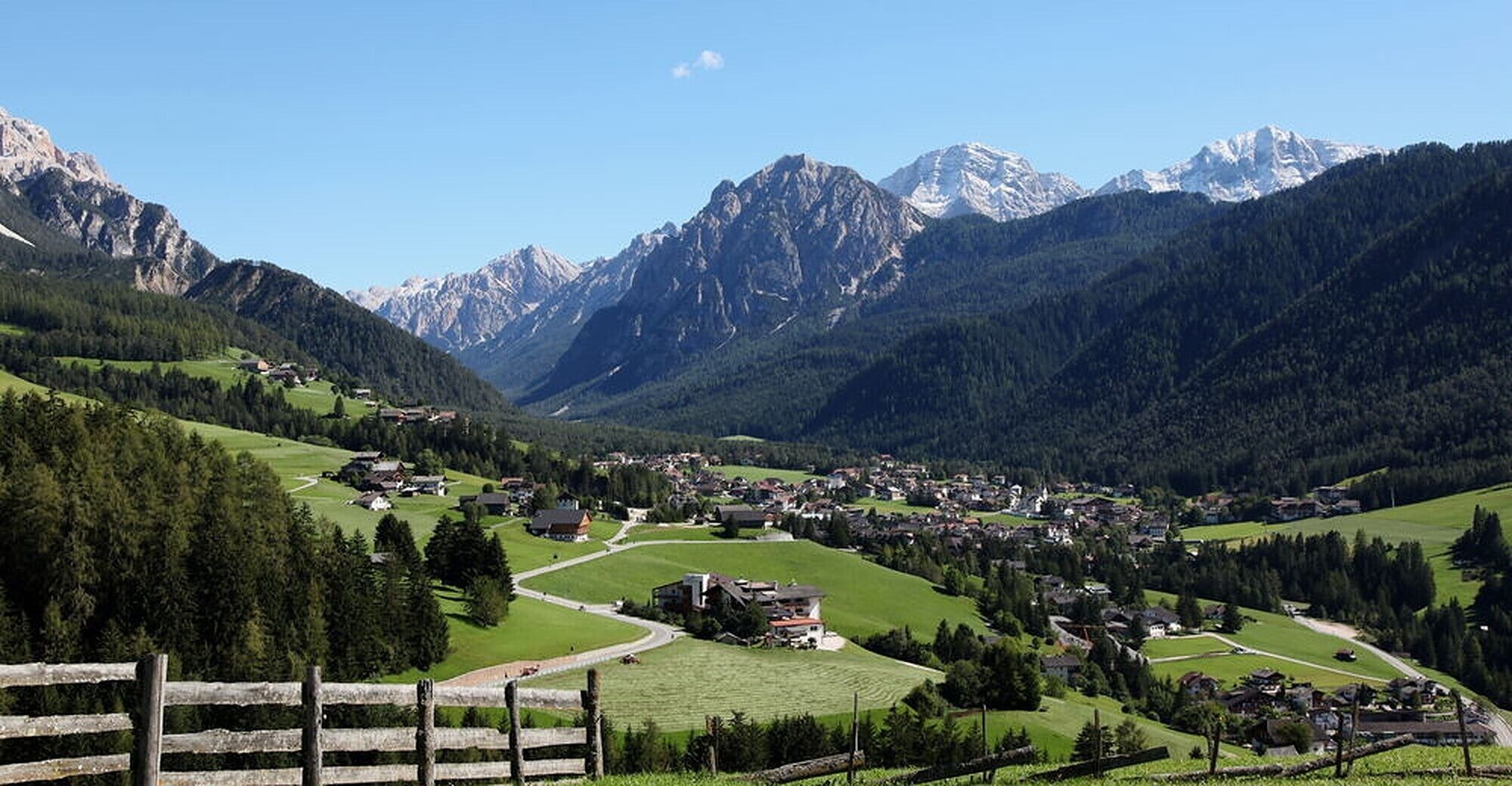 A mountain village with houses, a wooden fence, and snow-capped peaks in the background.