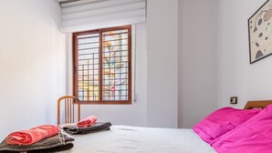 A bedroom with a bed, a window with a view of buildings, a chair, and a wall-mounted light switch. - Holiday Apartment in Alicante with Air Conditioning & WiFi (Alicante)