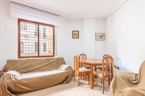 Una habitación con un sofá tapizado, una mesa de comedor de madera con sillas, una ventana con persianas y dos cuadros en la pared.