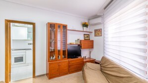 A living room with a wooden cabinet, a television, a sofa, and a window with blinds. - Holiday Apartment in Alicante with Air Conditioning & WiFi (Alicante)