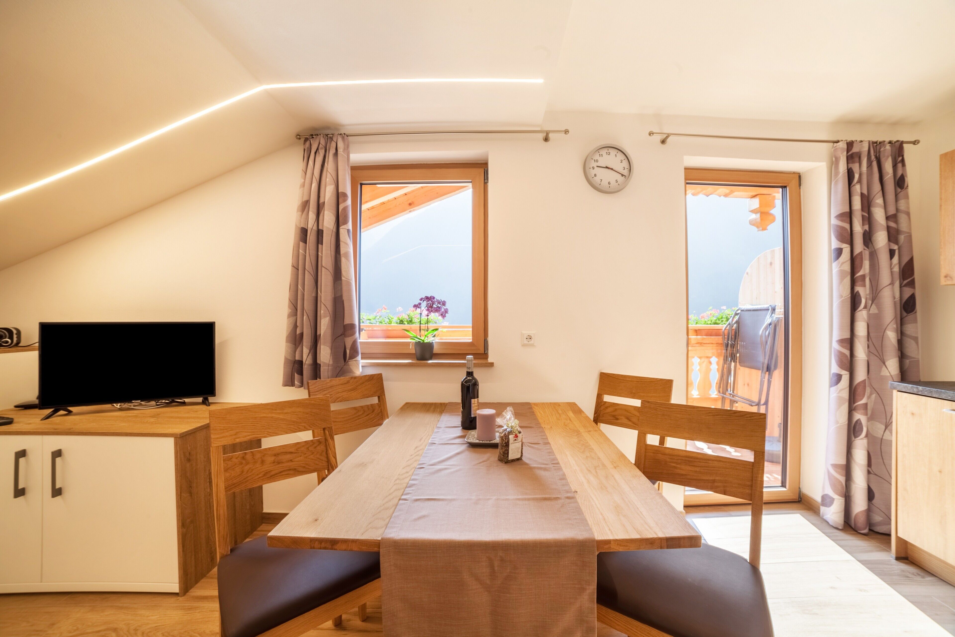 Un coin repas avec une table en bois, des chaises, un téléviseur et une fenêtre avec des rideaux.