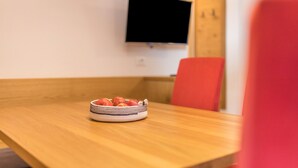 Ein Esstisch mit Obstschale, ein an der Wand befestigter Fernseher und ein roter Stuhl.